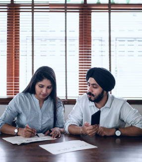 Two students collaborating at a table with documents, with one holding a pen and the other using a smartphone, sharing inspiring stories.