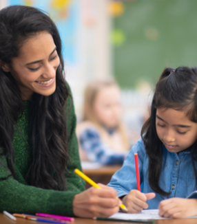 A smiling teacher, supported by the Neeyamo Foundation, assisting a young student with her work in a bright classroom setting.