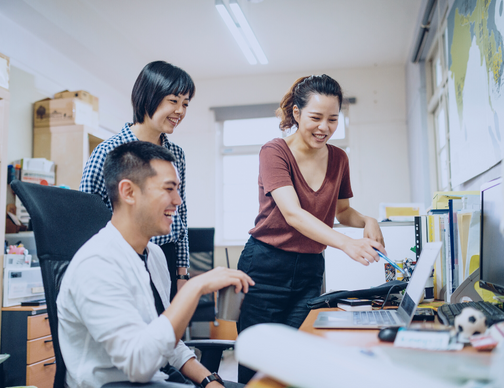 A diverse group of professionals brainstorm around a table, discussing global payroll solutions for mid-sized multinational corporations. Concepts like compliance, outsourcing, processing, and software are on a whiteboard behind them.