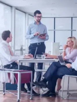 A business meeting in a modern office, with one man standing and using a tablet to discuss global payroll strategies, and two seated colleagues, a woman and a man, attentively listening.